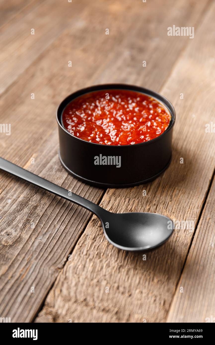 Red salmon caviar in an open black tin can and a black spoon on wooden background. Useful delicacy seafood, canned fish. Stock Photo