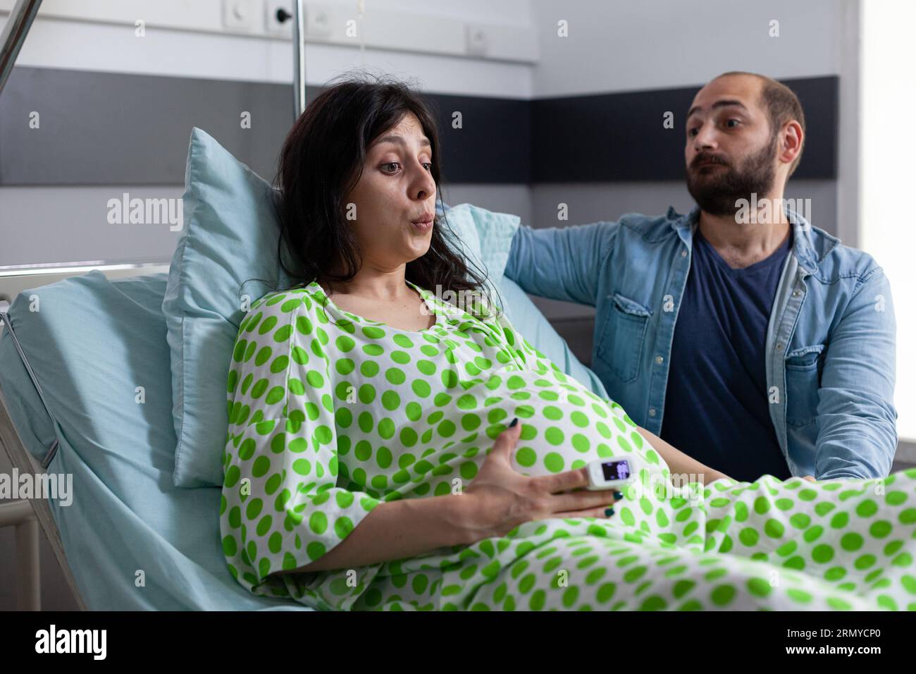 Pregnant woman doing breathing exercices, holding hands on belly while having painful contractions. Patient with pregnancy going into labor in hospital ward, husband standing beside her comforting. Stock Photo