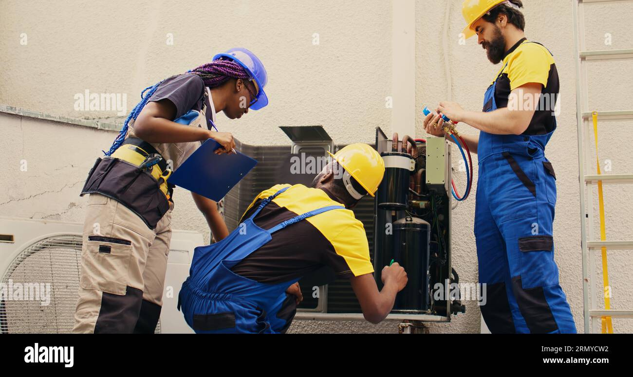 Expert repairmen cleaning layer of dirt and dust from air conditioner compressor coils while coworker uses manifold indicators to check for refrigerant leaks and read pressure in hvac system Stock Photo