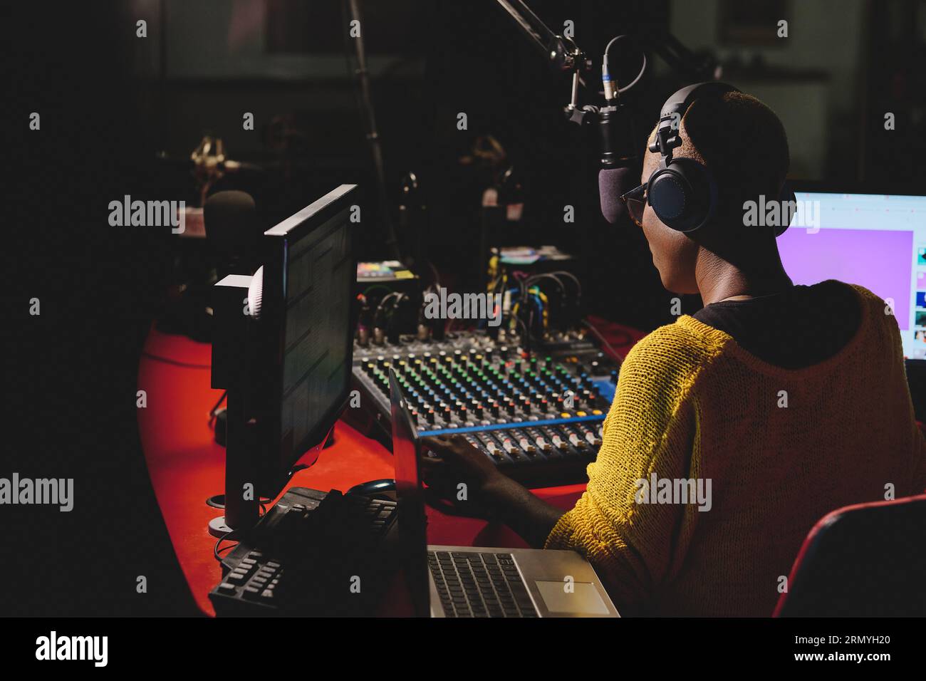 Back view of unrecognizable ethnic female in headphones using mixing console while sitting at table with laptop in dark broadcast studio and talking i Stock Photo