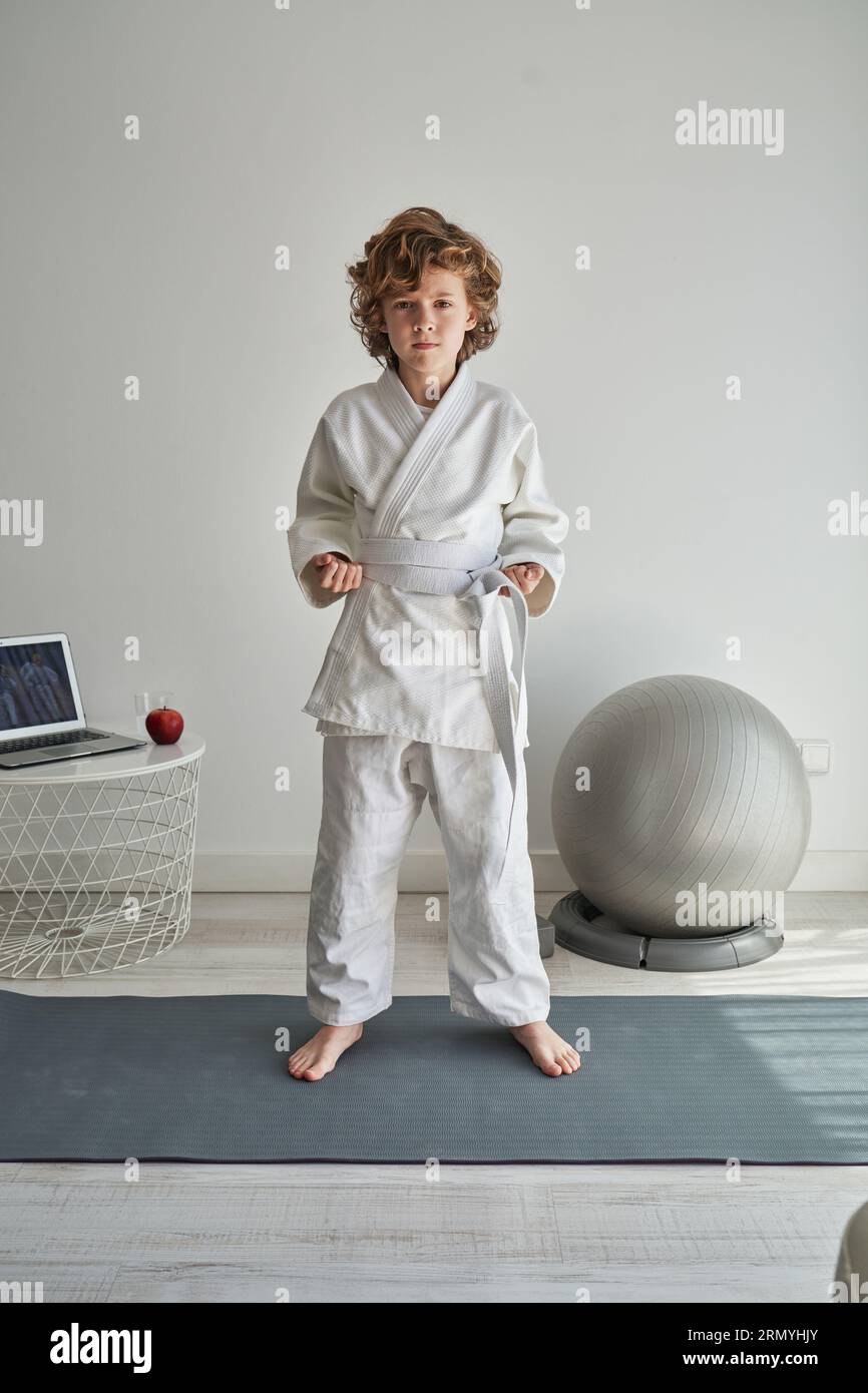 Full length of barefoot child in kimono and white belt standing on sports mat in pose and looking at camera while practicing judo at home Stock Photo
