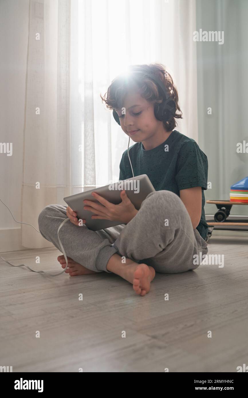 Full body of concentrated barefoot child in headphones sitting on floor with crossed legs near window in back lit and browsing internet via tablet dur Stock Photo