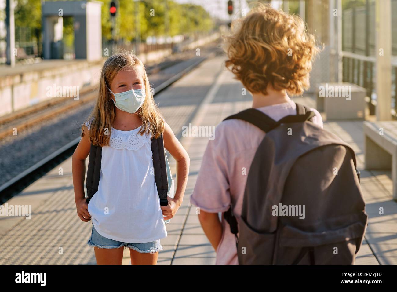 Preteen boy and girl in protective masks looking at each other and talking while waiting for train on platform during coronavirus epidemic Stock Photo