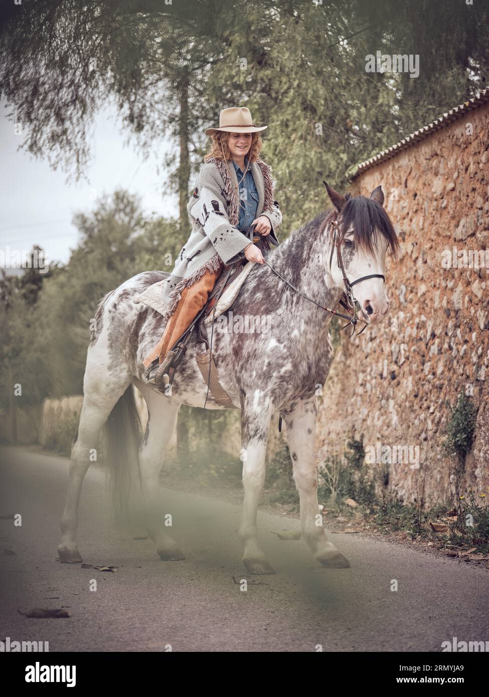 Smiling woman cowboy sitting on gray spotted horse and holding reins in hand while standing near fence Stock Photo
