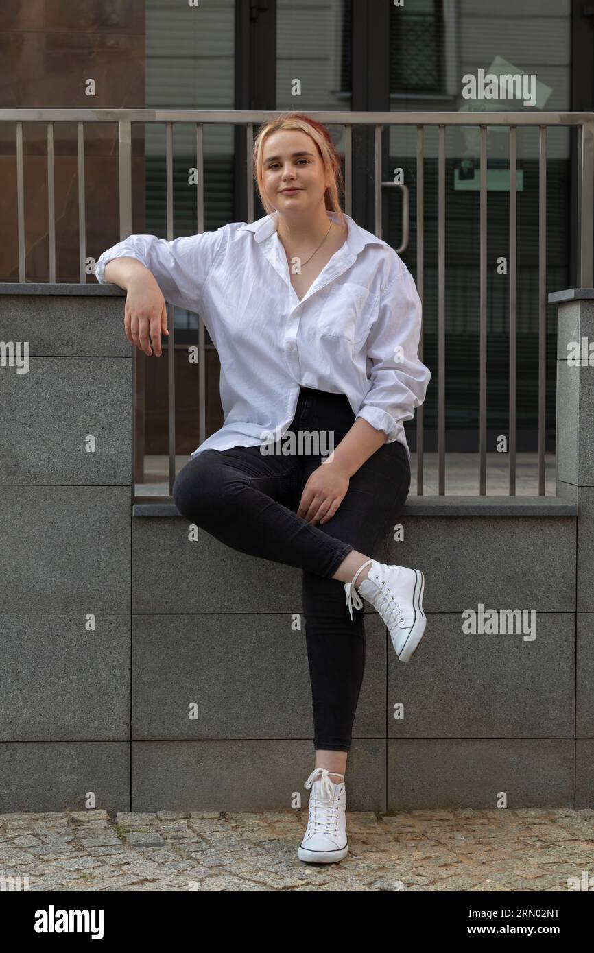 Full Length Young Real Beautiful Brown Haired Woman In Jeans And T-shirt Sitting On Stone Steps, Railing Outdoor. Carefree Dreamy Zoomer, Student Stock Photo
