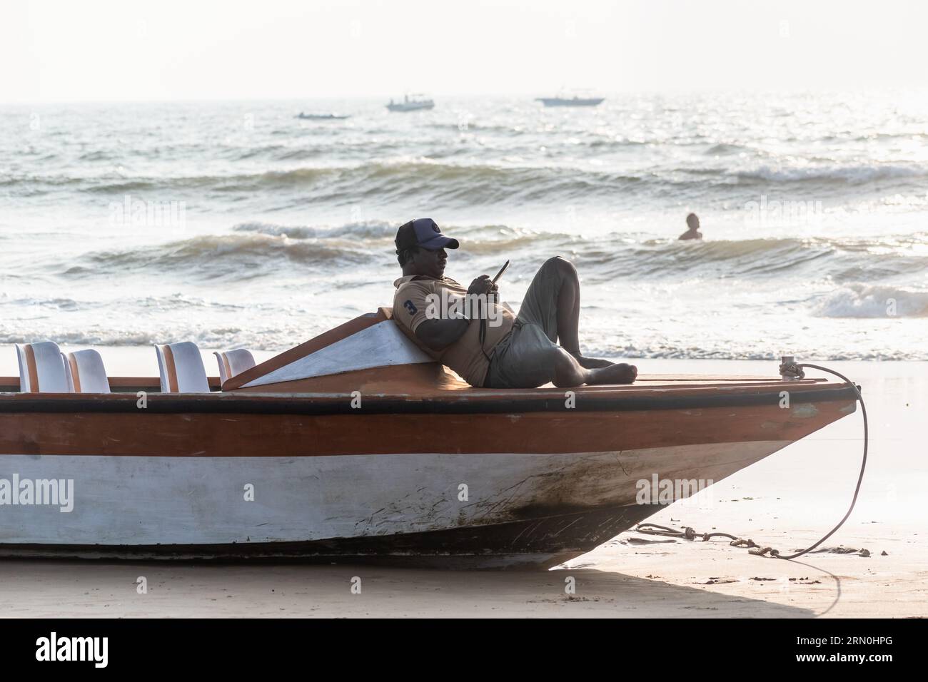 Calangute, Goa, India - January 2023: An Indian fisherman relaxing on a boat staring at his phone on a beach in Goa. Stock Photo