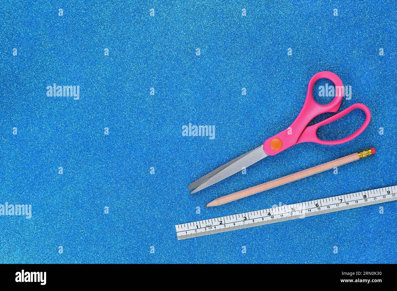 A flat, top view of an Arts and Crafts background with blue, sparkly, glitter paper, pink scissors, pencil and ruler with copy space to the left Stock Photo
