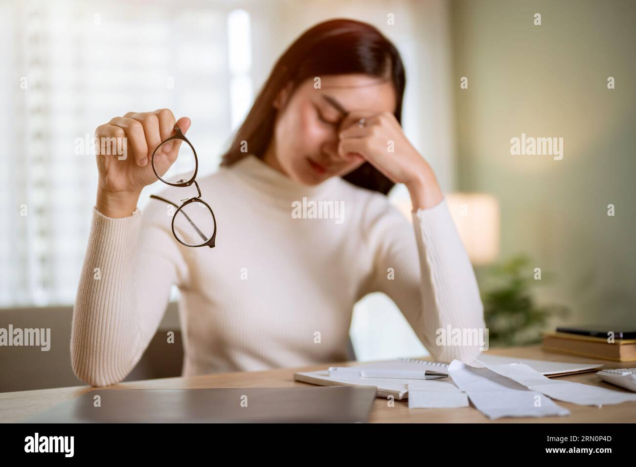 A stressed Asian woman is taking off her glasses while calculating her monthly household expenses and taxes, having headaches, and worried about finan Stock Photo
