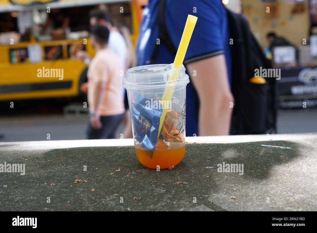 Plastic glass with orange drink, straw and small pieces of wrappers from chocolate lying on table as garbage. Stock Photo
