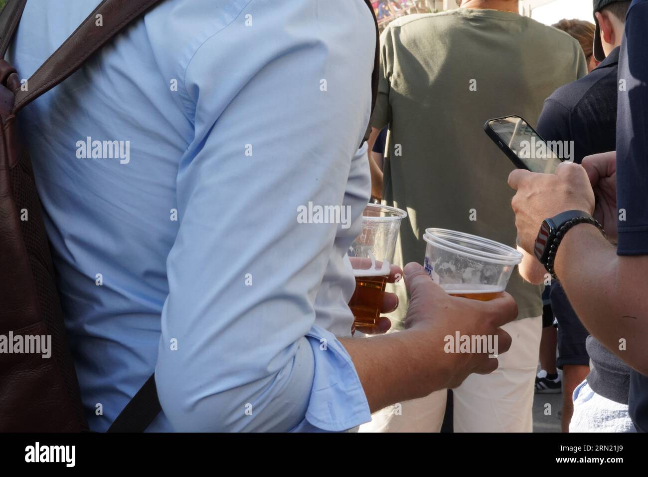 Young man with two plastic glasses with beer in each hand standing among other people during summer festival. Stock Photo