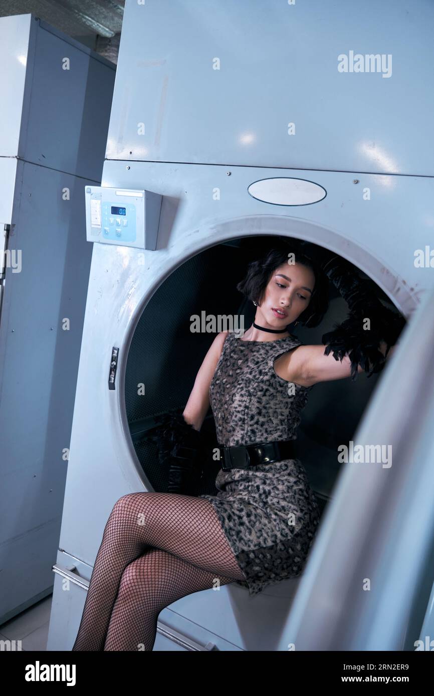 pretty young asian woman in dress and gloves posing in washing machine in self service laundry Stock Photo