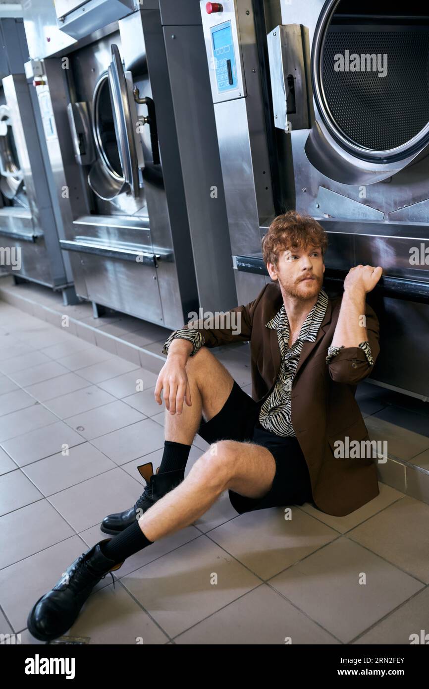 trendy redhead man in jacket posing near washing machine in public laundry Stock Photo
