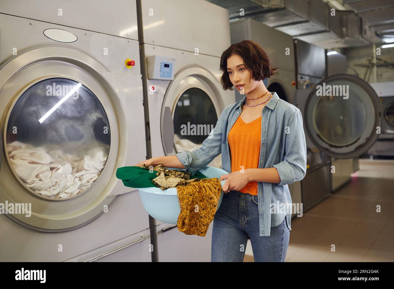 young asian woman holding basket with clothes near washing machines in public laundry Stock Photo