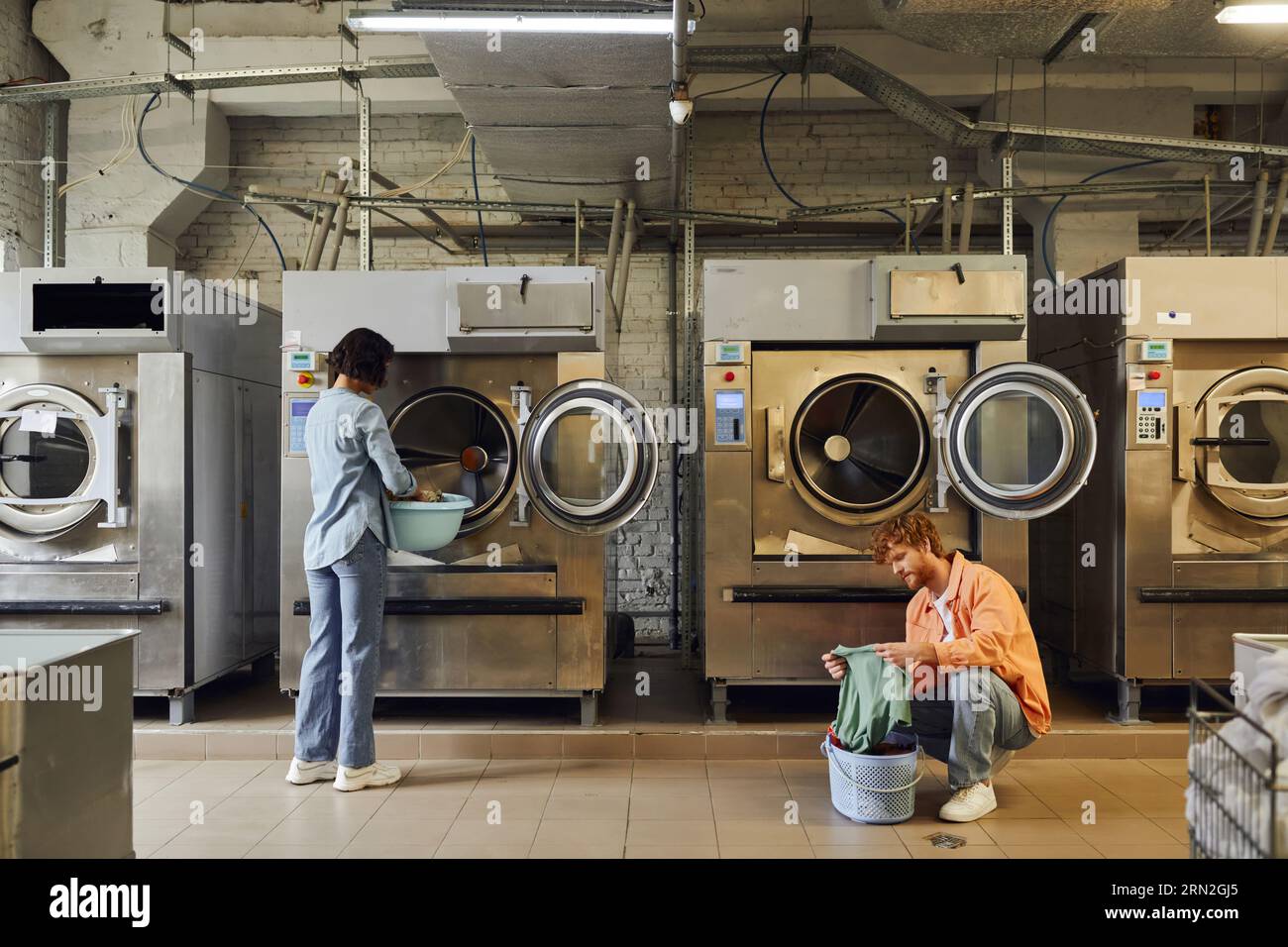 woman putting clothes in washing machine near boyfriend with basin in public laundry Stock Photo
