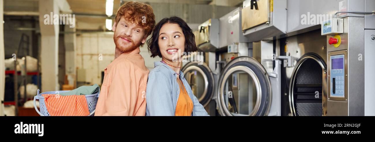 smiling man holding basket with clothes near asian girlfriend in coin laundry, banner Stock Photo