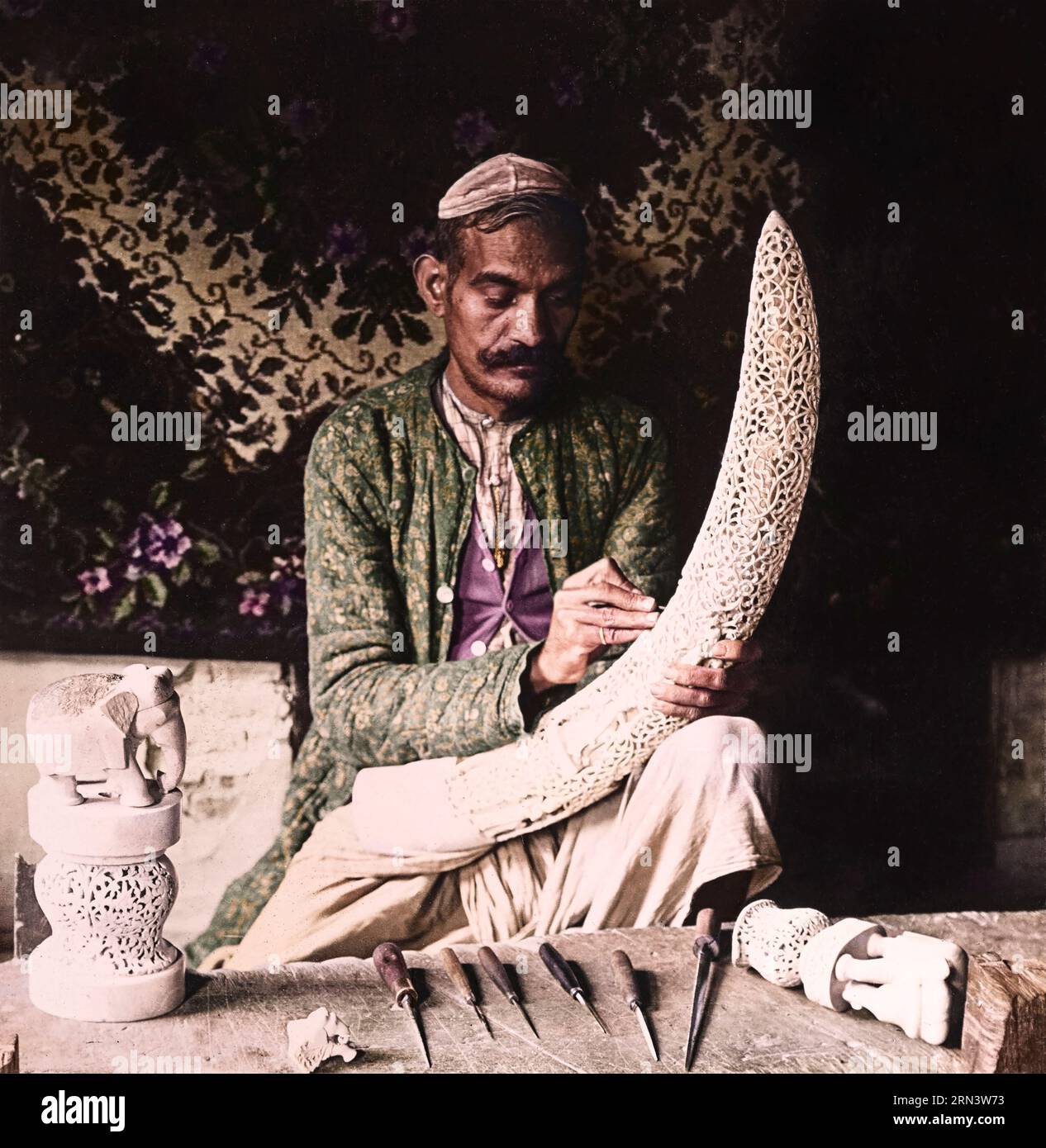 An Indian artist carving an elephant's tusk, in one of the workshops of Delhi, India. Year: 1907. By H.C. White Co. publishers. Stock Photo