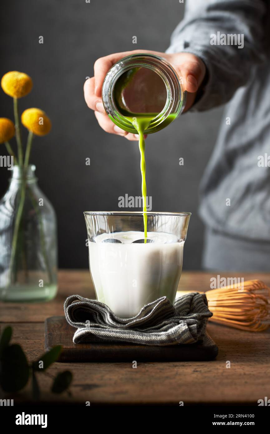 Cropped hand pouring shot of matcha to a glass of milk to prepare matcha latte. Wooden table and grey background. Living room or dining room setting Stock Photo