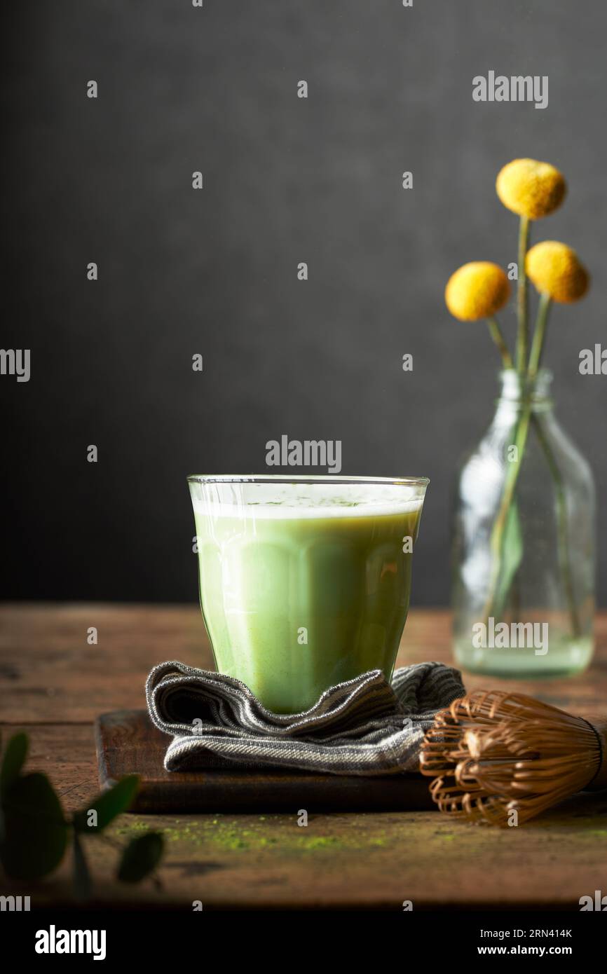 A glass of matcha latte on a rustic wooden table and grey background. Indoors set up. There are flowers, napkin, cutting board and bamboo whisk. Stock Photo
