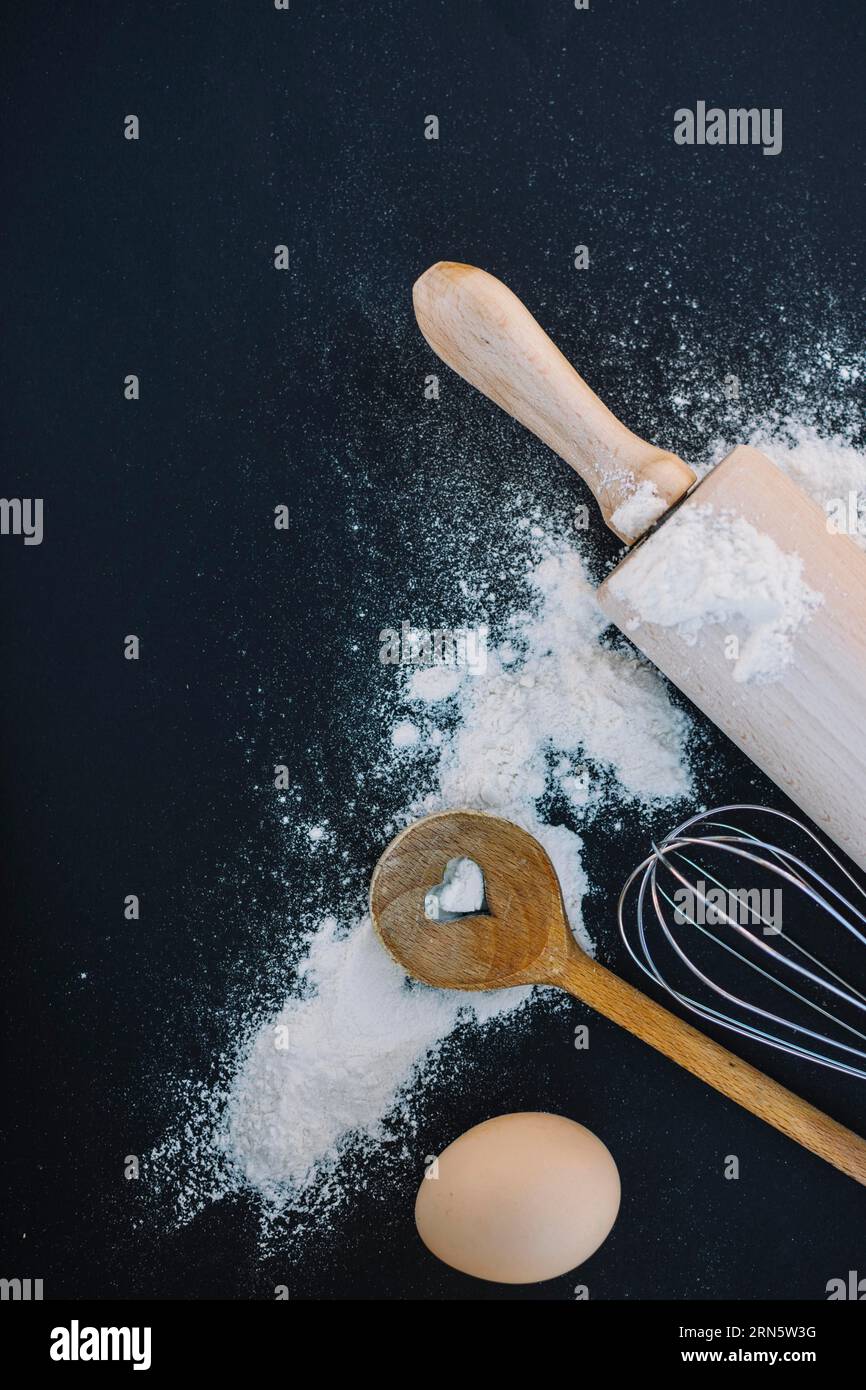 Top view heart shape spoon whisk flour egg rolling pin black background ...
