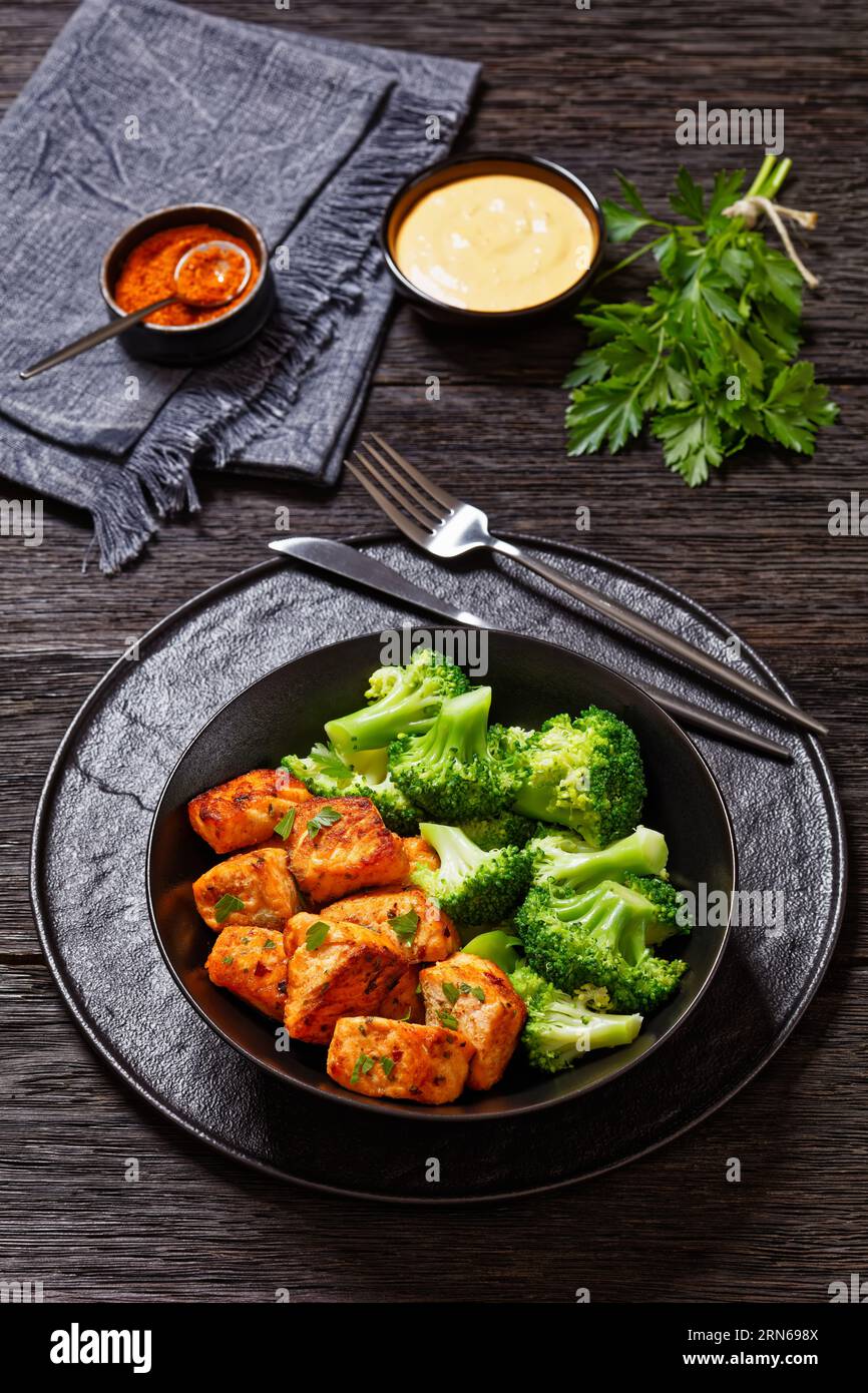 baked salmon chunks with steamed broccoli florets in black bowl on dark wooden table with sriracha mayo sauce, vertical view from above Stock Photo
