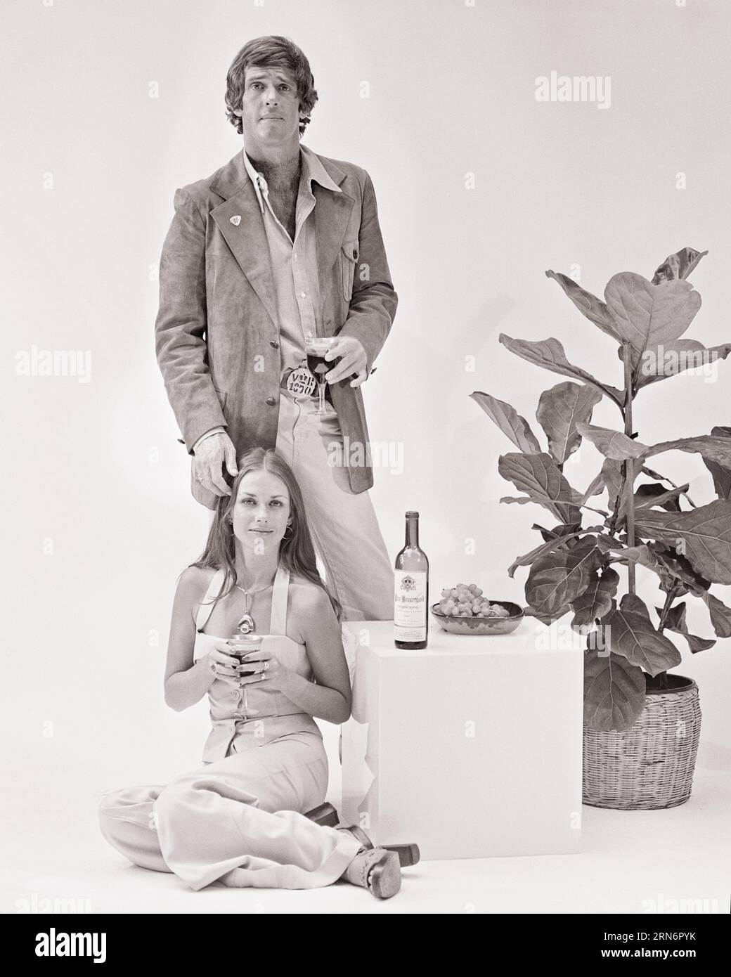 1970s FASHIONABLE COUPLE MAN STANDING WOMAN SITTING CUBE TABLE WITH WINE BOTTLE BOTH HOLDING GLASSES OF WINE LOOKING AT CAMERA - f13393 HAR001 HARS EYE CONTACT CUBE BEVERAGE STYLES JUMPSUIT FLUID HAIRSTYLE GRAPES HOUSE PLANT LIQUOR STYLISH SPIRITS BOOZE FASHIONS MID-ADULT MID-ADULT MAN MID-ADULT WOMAN SUEDE BEVERAGES BLACK AND WHITE CAUCASIAN ETHNICITY HAR001 OLD FASHIONED Stock Photo