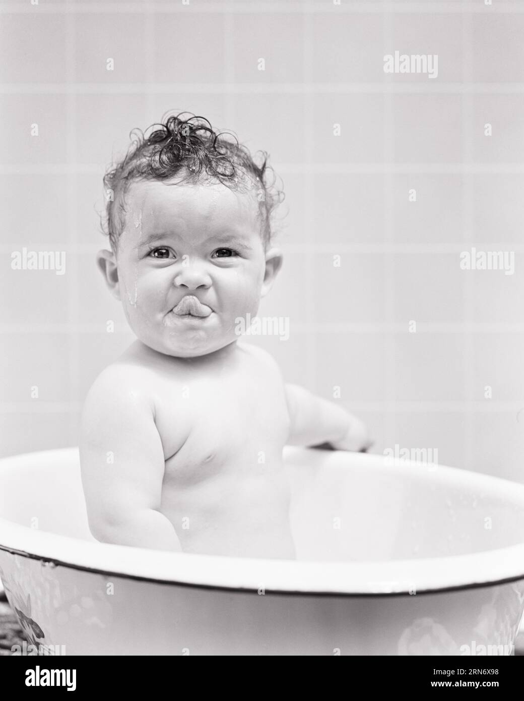 1940s SMILING BABY GIRL SITTING IN BATHTUB LOOKING AT CAMERA TONGUE LICKING TOP LIP  - b8783 HAR001 HARS BRUNETTE HUMOROUS LICKING EXCITEMENT COMICAL COMEDY STICKING OUT TONGUE PLEASANT AGREEABLE CHARMING COOPERATION GROWTH JUVENILES LIP LOVABLE PLEASING ADORABLE APPEALING BABY GIRL BLACK AND WHITE CAUCASIAN ETHNICITY HAR001 OLD FASHIONED Stock Photo