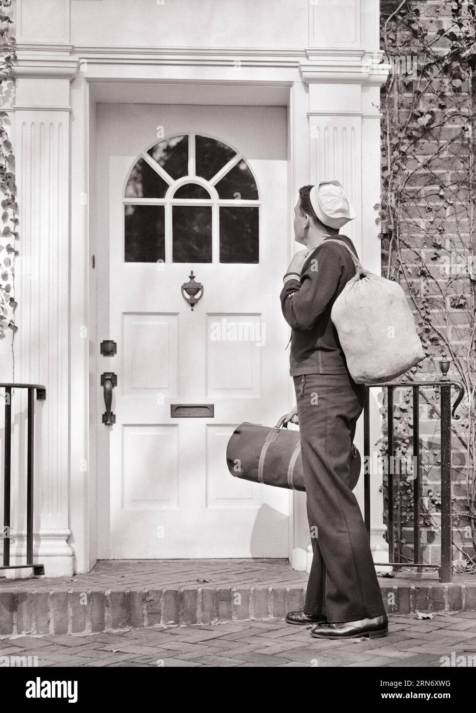 1940s BACK VIEW OF NAVY SAILOR IN UNIFORM HOLDING DUFFLE BAGS STANDING IN FRONT OF THE FRONT DOOR OF HOUSE COMING HOME - m2772 HAR001 HARS WW2 NAVY HOME LIFE UNITED STATES COPY SPACE FULL-LENGTH PERSONS UNITED STATES OF AMERICA MALES FORCE LEAVE B&W NORTH AMERICA COMING FREEDOM NORTH AMERICAN GLOBAL NAVAL EXCITEMENT EXTERIOR POWERFUL WORLD WARS PRIDE WORLD WAR WORLD WAR TWO REAR VIEW WORLD WAR II POLITICS UNIFORMS CONNECTION FORCES CONCEPTUAL FROM BEHIND NAVIES VETERAN WORLD WAR 2 IN FRONT OF VET USN BACK VIEW MID-ADULT MID-ADULT MAN RETURN RETURNING BLACK AND WHITE CAUCASIAN ETHNICITY HAR001 Stock Photo