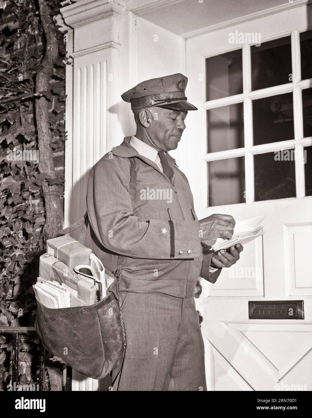 1960s PORTRAIT OF AFRICAN-AMERICAN MAILMAN USPS MAN WEARING UNIFORM DELIVERING MAIL TO DOOR CARRYING MAIL BAG ON HIS SHOULDER - p7193 HAR001 HARS PERSONS POSTAL UNITED STATES OF AMERICA MALES PROFESSION B&W NORTH AMERICA DELIVERING NORTH AMERICAN SKILL OCCUPATION SKILLS HIS CUSTOMER SERVICE AFRICAN-AMERICANS AFRICAN-AMERICAN CAREERS BLACK ETHNICITY LABOR EMPLOYMENT OCCUPATIONS MAILING CONCEPTUAL MAILMAN INFRASTRUCTURE EMPLOYEE USPS MID-ADULT MID-ADULT MAN UNITED STATES POSTAL SERVICE BLACK AND WHITE HAR001 LABORING OLD FASHIONED AFRICAN AMERICANS Stock Photo