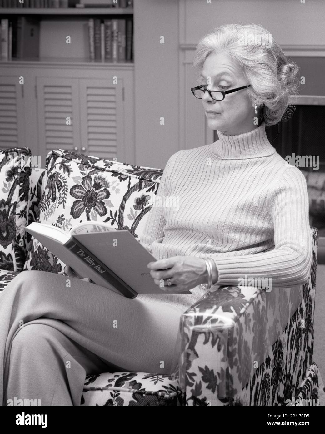 1970s FASHIONABLE MATURE WOMAN SITTING ON COUCH WEARING READER EYEGLASSES AND TURTLENECK SWEATER SLACKS READING A BOOK - r24217 HAR001 HARS PERSONS CONFIDENCE EYEGLASSES MIDDLE-AGED B&W MIDDLE-AGED WOMAN LEISURE LIVING ROOM AND RELAXED SLACKS ELDERS READER STYLISH READING GLASSES BLACK AND WHITE CAUCASIAN ETHNICITY HAR001 OLD FASHIONED TURTLENECK Stock Photo