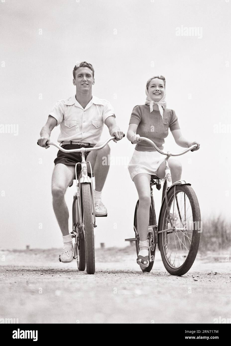 1940s SMILING YOUNG COUPLE MAN AND WOMAN WEARING SHORTS RIDING BEACH BIKES LOOKING AT CAMERA  - b17839 HAR001 HARS 1 FITNESS SHORTS HEALTHY YOUNG ADULT BALANCE TEAMWORK VACATION PLEASED JOY LIFESTYLE FEMALES MARRIED BIKING RURAL SPOUSE HUSBANDS HEALTHINESS COPY SPACE FRIENDSHIP FULL-LENGTH LADIES PHYSICAL FITNESS PERSONS INSPIRATION MALES SERENITY TOWARDS CONFIDENCE BICYCLES TRANSPORTATION B&W PARTNER BIKES EYE CONTACT FREEDOM GOALS TIME OFF ACTIVITY HAPPINESS PHYSICAL WELLNESS CHEERFUL ADVENTURE LEISURE STRENGTH TRIP AND GETAWAY EXCITEMENT LOW ANGLE RECREATION DIRECTION PRIDE OPPORTUNITY Stock Photo