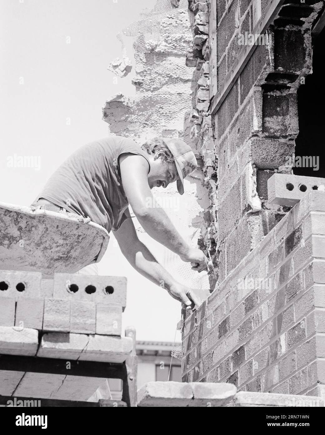 1970s MAN MASON BRICKLAYER WORKING TO REBUILD BRICK WALL OF BRICK HOUSE