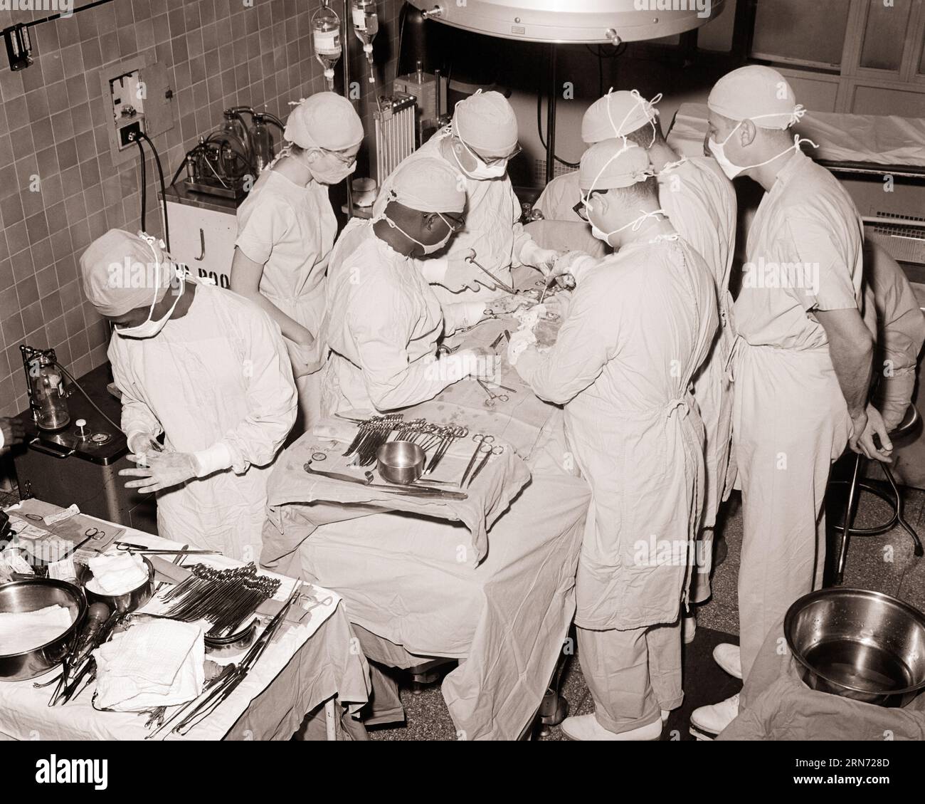1960s 1970s DOCTORS SURGEONS NURSES STERILE GOWNS CAPS MASKS PERFORMING SURGERY IN OPERATING ROOM OF HOSPITAL - m8451 HAR001 HARS RISK CONFIDENCE B&W HEALTHCARE PERFORMING SURGERY WIDE ANGLE OPERATING PREVENTION HEAD AND SHOULDERS HIGH ANGLE STERILE STRENGTH HEALING AFRICAN-AMERICANS AFRICAN-AMERICAN DIAGNOSIS SURGICAL KNOWLEDGE SURGEONS BLACK ETHNICITY GOWNS OPERATING ROOM HEALTH CARE AUTHORITY IMPAIRMENT OCCUPATIONS SURGEON TREATMENT CONCEPTUAL CAPS HOSPITALS OPERATION STYLISH FACILITY FACILITIES OR COOPERATION BLACK AND WHITE CAUCASIAN ETHNICITY DISEASE HAR001 OLD FASHIONED Stock Photo