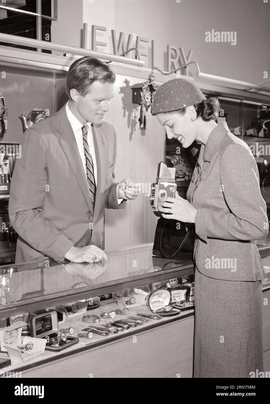 1950s STYLISH WOMAN SHOPPING AT GIFT COUNTER TALKING TO SALESMAN AS SHE HOLDS A TWIN LENS REFLEX CAMERA AS POSSIBLE PURCHASE - s7001 HAR001 HARS MALES GIFTS B&W LENS SKILL SUIT AND TIE ACTIVITY AMUSEMENT SELLING HAPPINESS WATCHES HOBBY CUSTOMER SERVICE INTEREST HOBBIES KNOWLEDGE PASTIME SHE INNOVATION PLEASURE OPPORTUNITY OCCUPATIONS PURCHASE STYLISH REFLEX HOLDS MID-ADULT MID-ADULT MAN MID-ADULT WOMAN POSSIBLE RELAXATION SALESMEN YOUNG ADULT MAN YOUNG ADULT WOMAN AMATEUR BLACK AND WHITE CAUCASIAN ETHNICITY ENJOYMENT HAR001 OLD FASHIONED Stock Photo