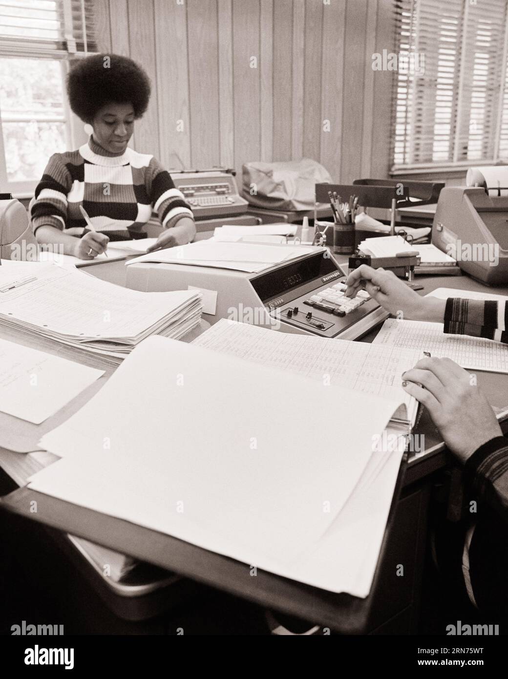 1970s AFRICAN AMERICAN WOMAN WITH AFRO HAIRSTYLE WORKING IN AN ACCOUNTING OFFICE CAUCASIAN HANDS USING AN ELECTRIC CALCULATOR  - o4052 HAR001 HARS B&W GOALS AFRICAN-AMERICANS AFRICAN-AMERICAN HAIRSTYLE LOW ANGLE ADDING MACHINE CLERKS BOOKKEEPER BLACK ETHNICITY OFFICE WORKER OCCUPATIONS USING GAL FRIDAY ACCOUNTING ADMINISTRATOR CALCULATORS SECRETARIES SUPPORT AMANUENSIS BOOKKEEPING COOPERATION MID-ADULT MID-ADULT WOMAN TOGETHERNESS BLACK AND WHITE CAUCASIAN ETHNICITY CLERICAL HANDS ONLY HAR001 OLD FASHIONED AFRICAN AMERICANS Stock Photo