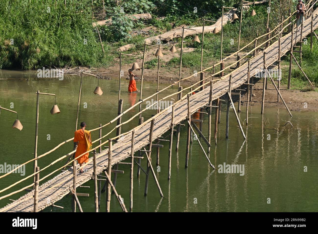 Young buddhist monk walking on a bamboo bridge on a river in Luang Prabang, Laos Stock Photo