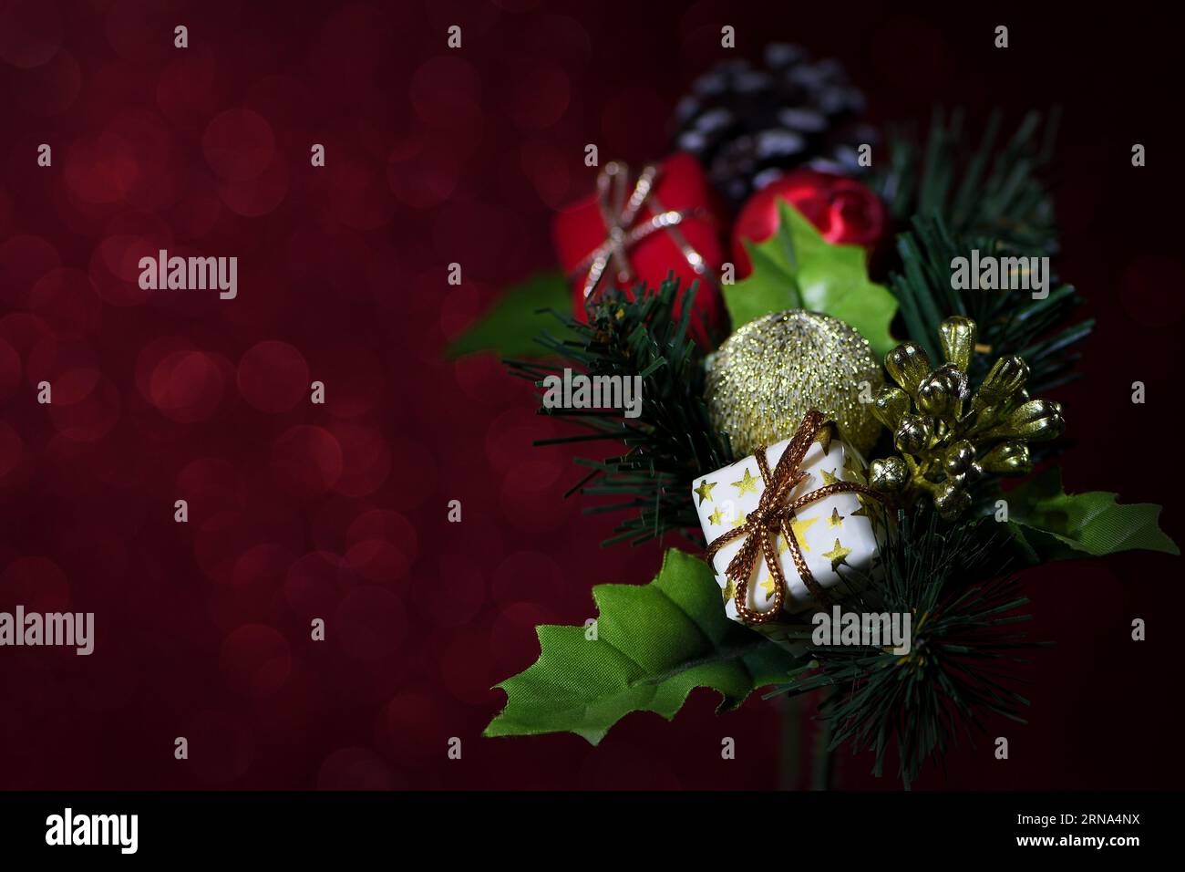 A rich, mood lit, colourful Christmas decoration on a sparkly red background with pine cones, presents, baubles and holly leaves with a shallow focus Stock Photo