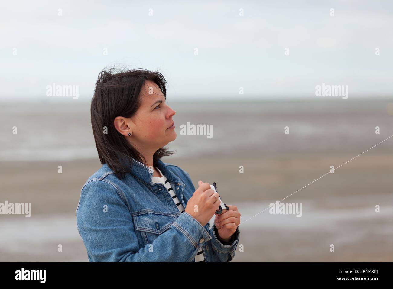 A middle-aged woman dressed in a denim jacket flies a kite on the beach at low tide. Bad weather during summer vacation Stock Photo