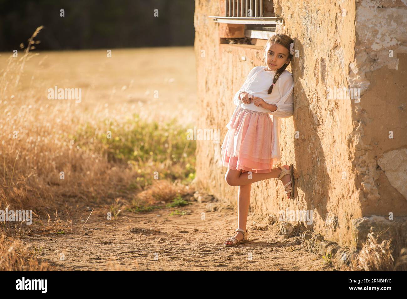little girl in pink skirt leaning against rustic wall at golden hour ...