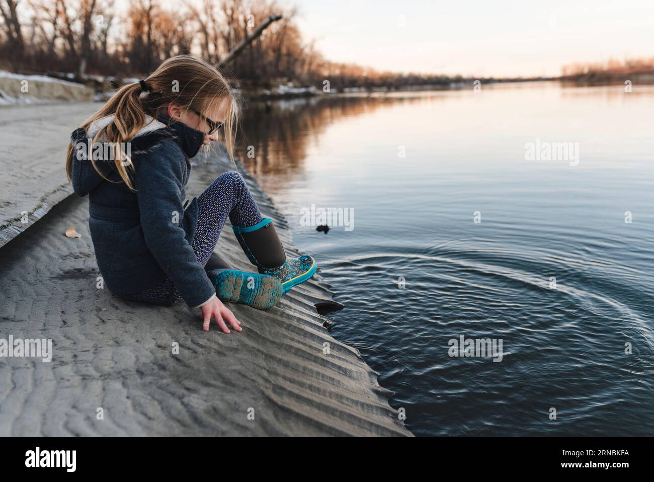 Girl wearing boots sits at edge of Missouri riverbank during fall Stock Photo