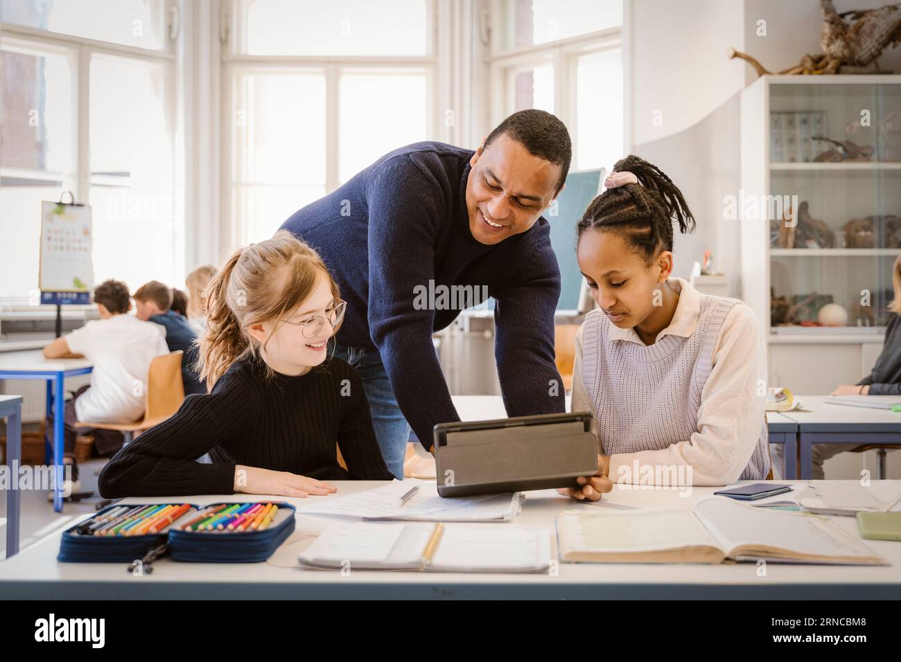Smiling male teacher doing E-learning with female students in classroom ...