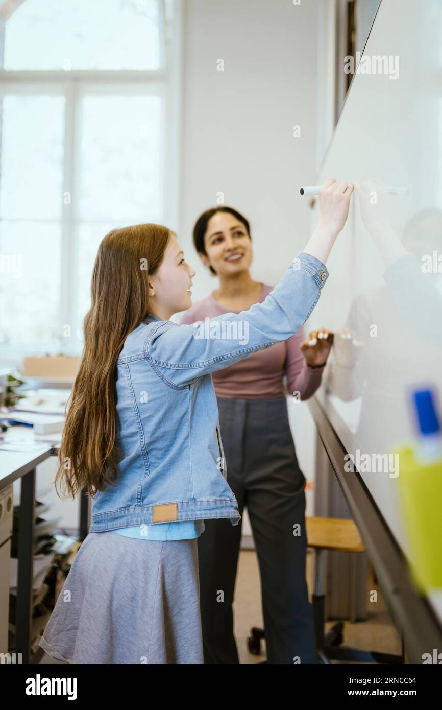 Side view of schoolgirl writing on whiteboard with felt tip pen by teacher in classroom Stock Photo