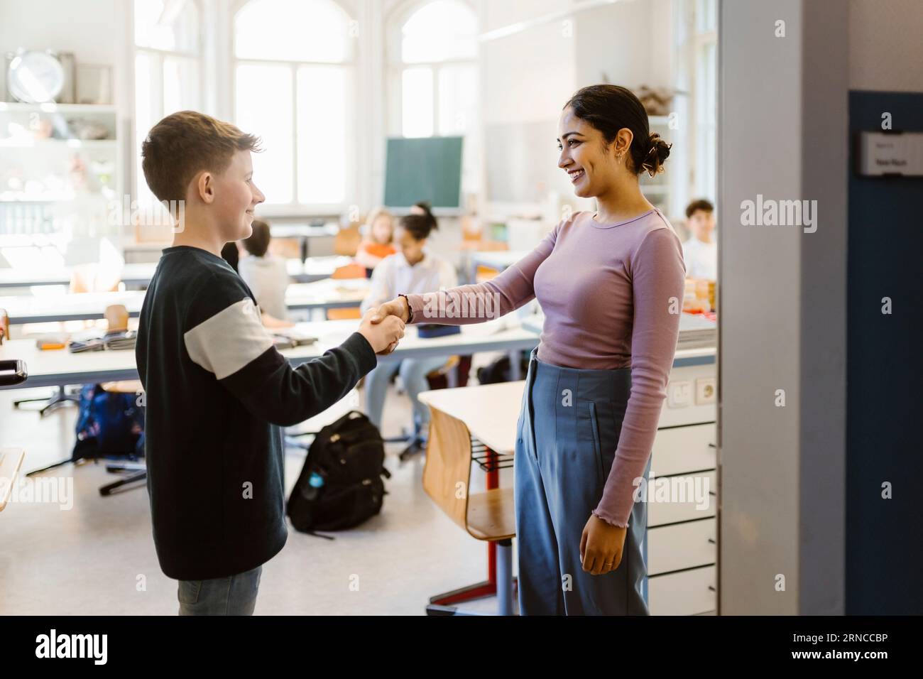 Smiling teacher shaking hand with schoolboy standing in classroom Stock Photo