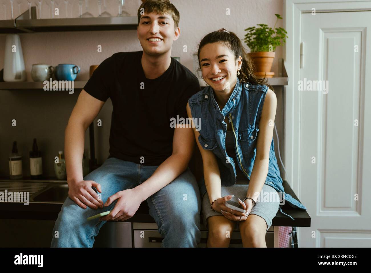 Portrait of smiling young friends sitting together on kitchen counter ...