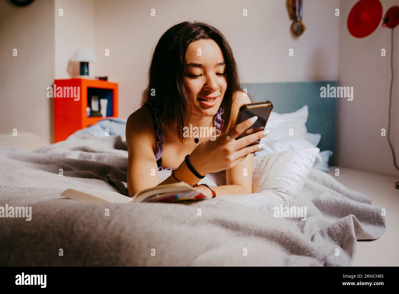 Smiling young woman using smart phone while lying on bed at home Stock Photo