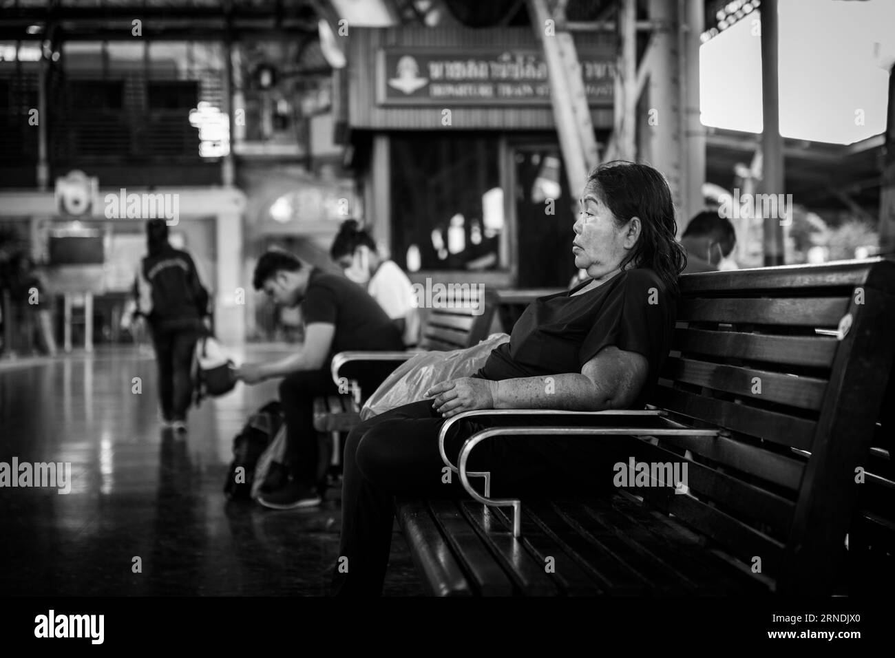In this black and white photograph, an elderly Thai woman sits contemplatively on a waiting bench at Hua Lamphong Railway Station in Bangkok, Thailand Stock Photo