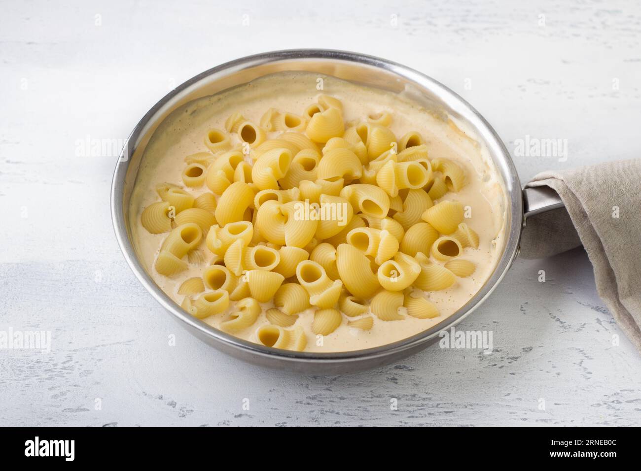 Cooking mac and cheese, American pasta. Adding cooked pasta to cheese sauce in a frying pan on a light gray background. Cooking stage Stock Photo