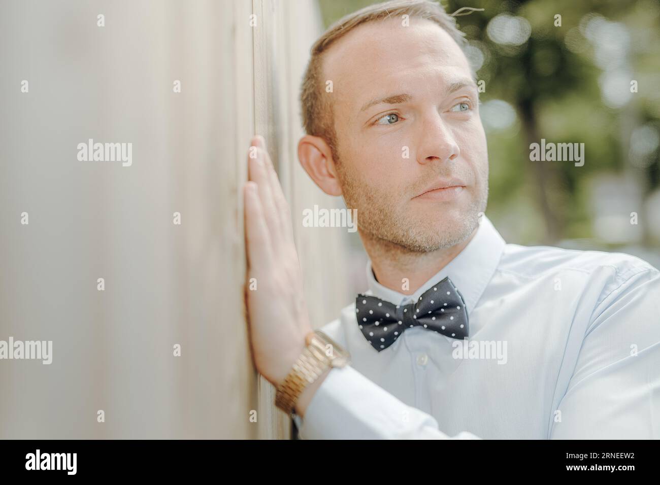 adult man with dotted bow tie has hand on wooden fence and lean with head to wall while look to side Stock Photo