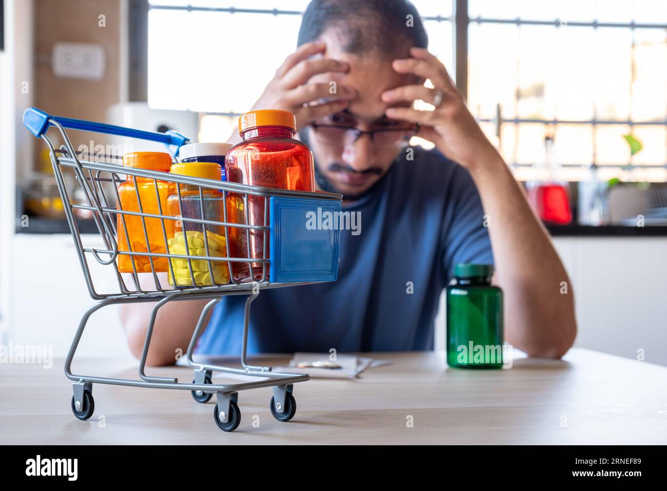 man sitting in the kitchen feeling frustrated due inflation and increase of daily needs supplies for house with shopping cart in front of him holding Stock Photo