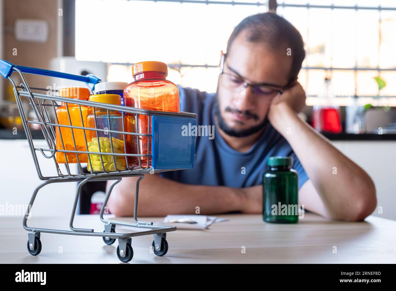 man sitting in the kitchen feeling frustrated due inflation and increase of daily needs supplies for house with shopping cart in front of him holding Stock Photo
