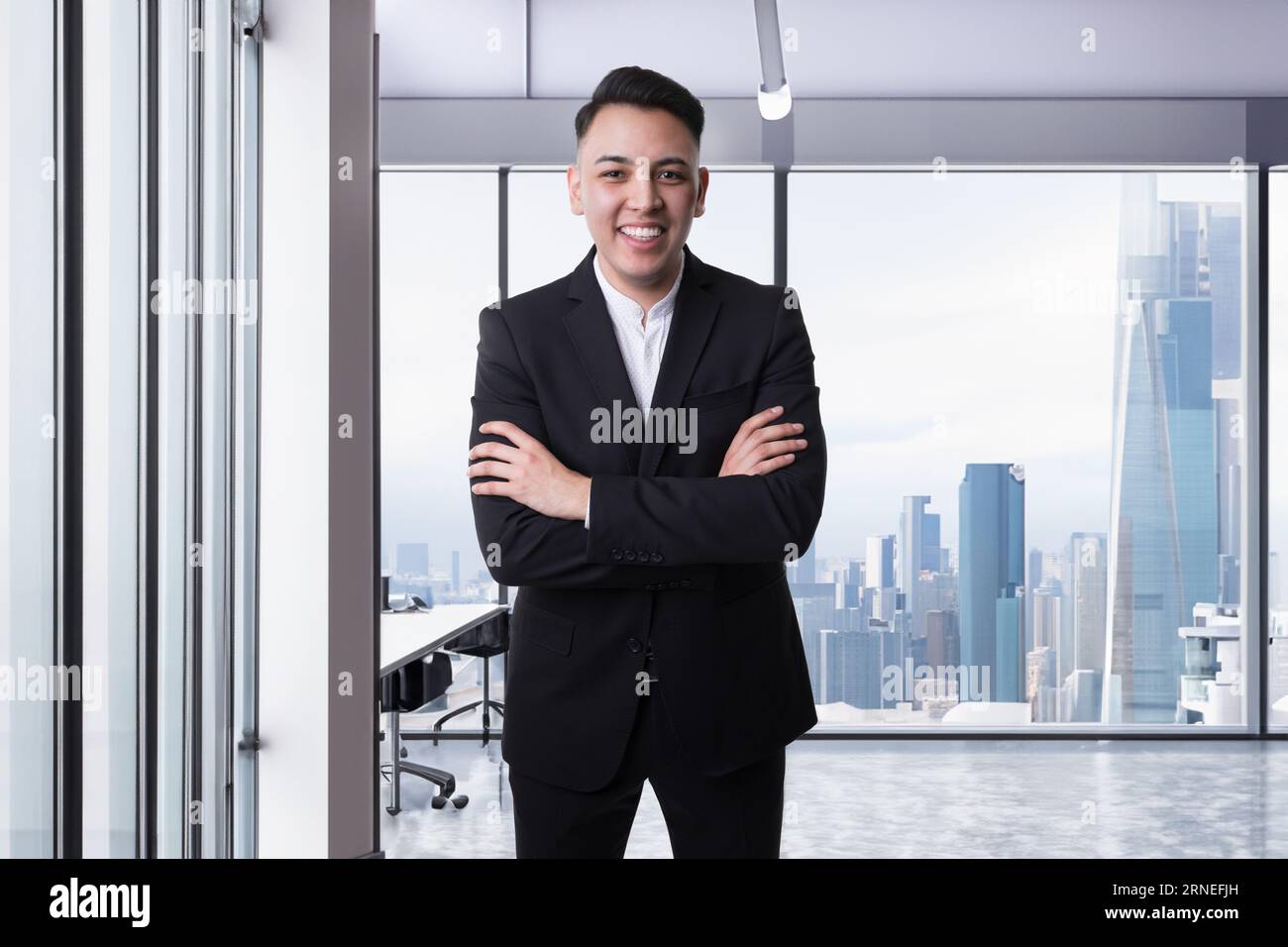 mexican young business man stand with crossed arms in front of big window with city skyline in background while he smile and is happy about his succes Stock Photo